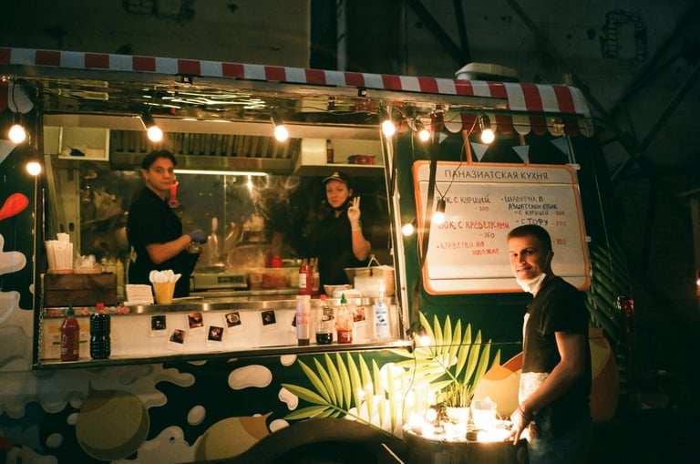Nighttime outdoor food or cosmetics vendor booth with striped awning, illuminated by string lights, staff inside counter, and merchandise displayed with tropical plants.