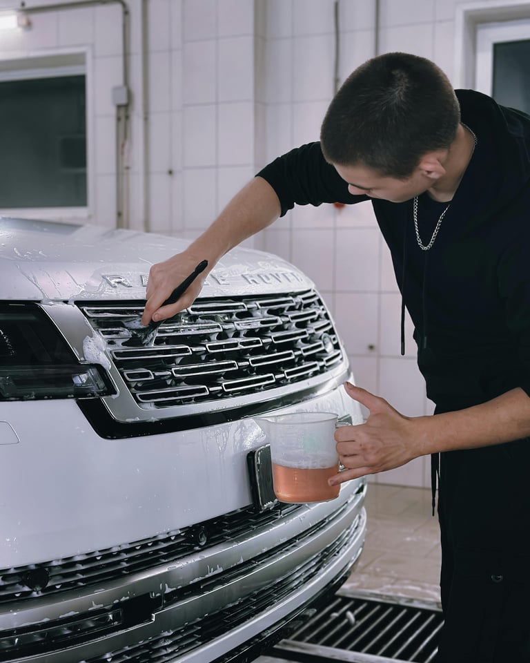 Man in black shirt applying protective film to the front grille and headlight area of a white SUV in a garage