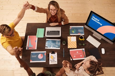 Three people working collaboratively around a desk filled with laptops, notebooks, and design materials, viewed from above.